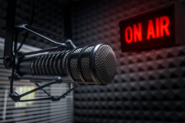 The image shows a close-up of a professional microphone in a recording studio. The microphone is mounted on a stand with a shock mount, and it has a metallic mesh grille. In the background, there is a red illuminated sign that reads "ON AIR," indicating that a live broadcast or recording session is in progress. The walls of the studio are covered with soundproofing foam panels, which help to reduce echo and noise. The overall atmosphere suggests a radio or podcasting environment.